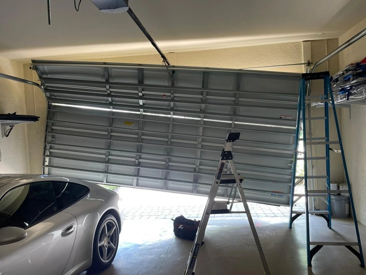 Garage door hanging at an angle over a parked car, with two ladders set up in the opening