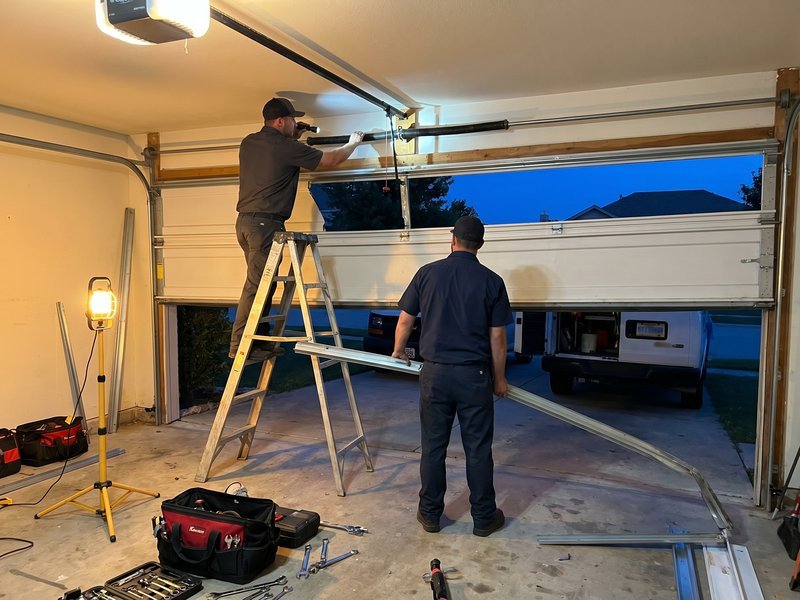 Two technicians working by lamp light on an open garage door at dusk, one on a stepladder